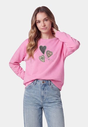 Giovane donna con lunghi capelli mossi che indossa una felpa rosa con tre cuori di paillettes e jeans a vita alta azzurri chiari.