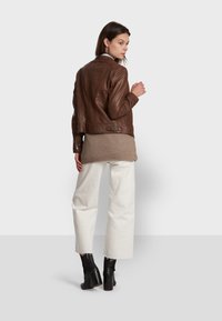 Brown leather jacket with a fitted design, side zippers, and a belted waist, worn over a beige knit top and paired with white wide-leg pants.