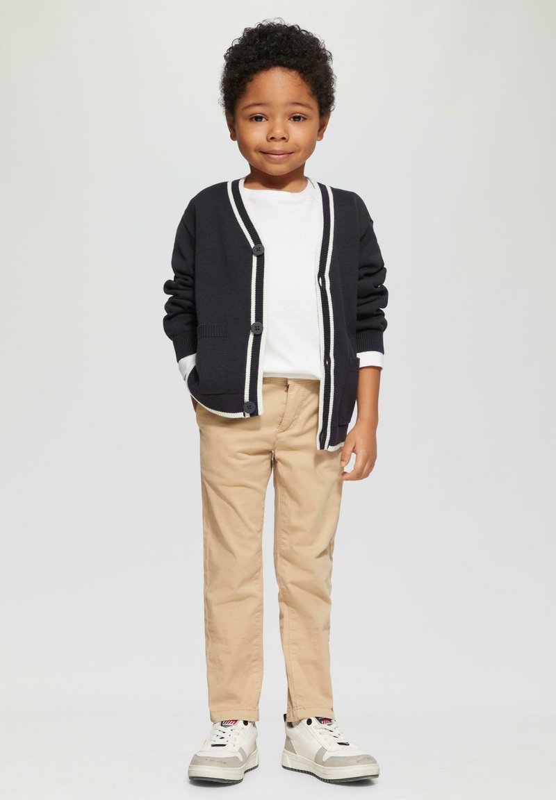 Marineblauwe cardigan met witte biezen en knopen, witte t-shirt, beige broek en witte sneakers met grijze accenten, geschikt voor kinderen.