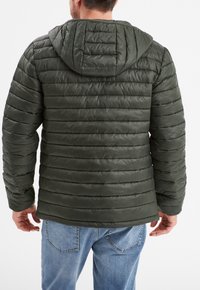 Chaqueta acolchada en verde oscuro con capucha, con líneas acolchadas, puños elásticos y una textura suave y brillante. Llevada sobre jeans azules.