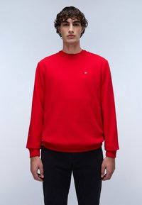 Sudadera roja con cuello y puños de canalé, que presenta un pequeño logo. Combinada con pantalones negros, se muestra sobre un fondo neutro.