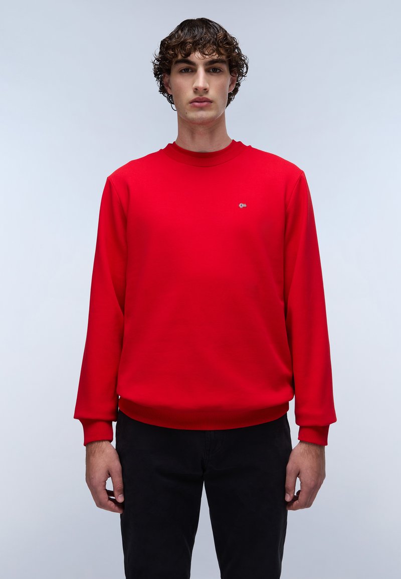 Sudadera roja con cuello y puños de canalé, que presenta un pequeño logo. Combinada con pantalones negros, se muestra sobre un fondo neutro.