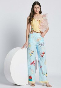 Rumena crop majica z nabranimi prozornimi detajli; svetlo modre hlače z različnimi barvnimi vzorci; model pozira poleg krožne dekoracije.