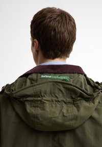 Άτομο με κοντά καστανά μαλλιά που φοράει μια χακί πράσινη ζακέτα με μπορντό γιακά που δείχνει μια ετικέτα "Barbour αγαπάει τον Paul Smith".