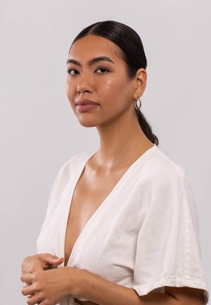 Model mit langen, dunklen Haaren in einem weißen V-Ausschnitt-Oberteil. Sie trägt goldene Creolen und hat einen glatten, strahlenden Teint.