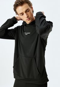 Sudadera negra con capucha y mangas largas, que presenta un bolsillo tipo canguro y un gráfico en texto "FORGE STRENGTH FOCUS" en el pecho. Textura de tela suave.