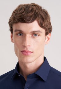 Camicia blu navy con colletto appuntito, realizzata in tessuto liscio. Il soggetto ha capelli corti e mossi e un'espressione neutra.