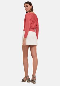 Koraalkleurige gebreide trui met brede halslijn, lange mouwen en geplooide achterkant, gecombineerd met een witte mini-jurk en geblokte sandalen met hak.