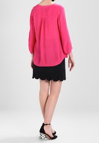 Donna che indossa una blusa a maniche lunghe di un brillante rosa, una mini gonna nera con bordi ondulati e tacchi a stiletto neri con tacco squadrato argentato, in piedi di spalle.
