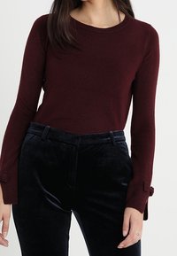 Burgundy knitted sweater with long sleeves, featuring a crew neckline and textured fabric. Paired with navy velvet trousers with a smooth finish.