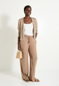 Cardigan brillante beige claro sobre una camiseta blanca, combinado con pantalones sueltos beige, y sosteniendo un pequeño bolso de mimbre. Las sandalias completan el look.