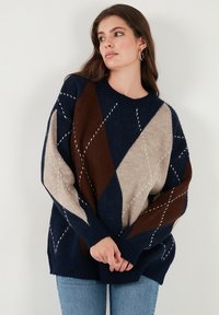 Knit sweater featuring a diamond pattern in navy, beige, and brown hues. Loose fit with ribbed neckline and cuffs. Textured fabric.
