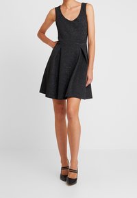 Svart ärmlös klänning med v-ringning, strukturerad bodice och utsvängd kjol med veck. Slät textur med en subtil glans.