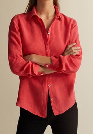 Personne les bras croisés portant une chemise corail à manches longues boutonnée et un pantalon noir sur un fond beige uni.