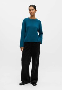 Teal, ronde-hals tröja med långa ärmar, parat med svarta, vida corduroybyxor. Tyget ser slätt och mjukt ut.