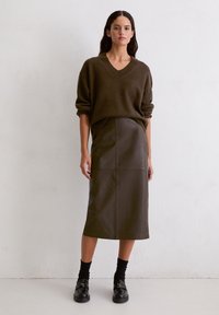 Brauner gestrickter Pullover mit V-Ausschnitt, kombiniert mit einem dunkelgrünen Leder-Midirock. Das Modell trägt schwarze Loafers und gerippte schwarze Socken.