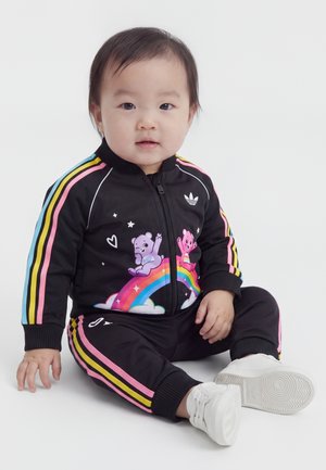 Tout-petit assis portant un survêtement noir avec des rayures colorées et des ours dessinés sur un arc-en-ciel, assorti à des baskets blanches.