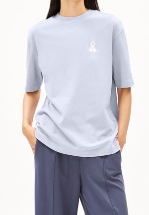Personne portant une chemise bleu clair à manches courtes avec un petit logo blanc sur la poitrine et un pantalon ample bleu foncé, mains dans les poches.