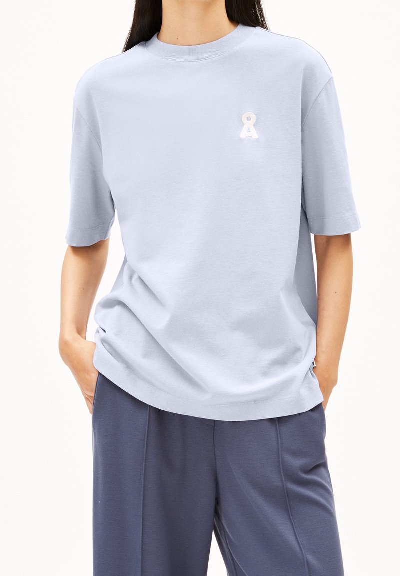 Personne portant une chemise bleu clair à manches courtes avec un petit logo blanc sur la poitrine et un pantalon ample bleu foncé, mains dans les poches.