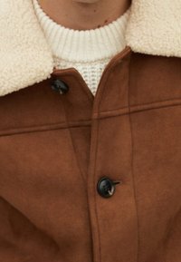 Brun ruskindsjakke med en lys fleecekrave, der har mørke knapper og subtile stingdetaljer. Båret over en lysstrikket sweater.