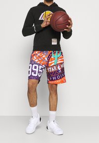Hoodie noir avec logo, short multicolore avec imprimés graphiques, comprenant des chiffres et un design NBA, et baskets blanches. Tenant un ballon de basket.