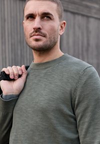 Homme aux cheveux courts et barbe naissante portant un pull vert, tenant une sangle noire sur l'épaule, regardant pensivement devant lui.