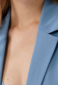 Světle modrý blejzr s klopovým límcem, vyrobený z hladkého materiálu s minimalistickým designem, částečně odhalující výstřih.