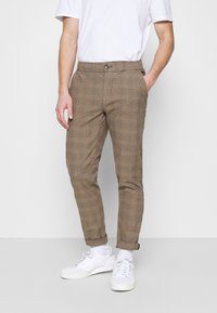 Pantalons à carreaux marron avec une coupe décontractée et une taille élastique. Comprend une fermeture à bouton et des ourlets à revers. Associés à des baskets blanches.