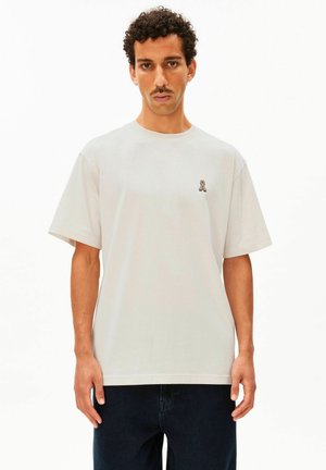 Jonge man met krullend haar en snor, die een effen wit T-shirt en donkere broek draagt, staand tegen een witte achtergrond.