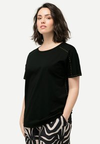 Camiseta negra de manga corta con acentos de tachuelas metálicas en los hombros. Tela suave y lisa, corte relajado, combinada con pantalones estampados en blanco y negro.