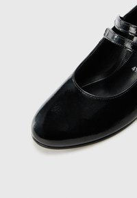 Chaussure en cuir verni noir avec un bout arrondi et une finition lisse. Présente deux fines brides traversant l'arche pour un ajustement sécurisé.