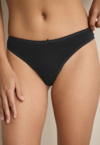 Perizoma in cotone nero con una texture liscia, caratterizzato da un delicato bordo in pizzo lungo la vita e un piccolo fiocco al centro.