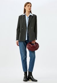 Blazer gris de doble botonadura, camisa azul clara, vaqueros azules, botines negros de tobillo, sosteniendo un bolso redondo de cuero burdeos. Diseño simple y a medida.