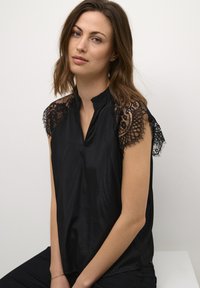 Blusa nera con maniche corte e dettagli in pizzo, colletto alto e una texture liscia, leggermente lucida. Senza motivi o decorazioni distintive.