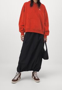 Sudadera con capucha roja con bolsillo canguro y logo, combinada con una falda maxi negra y zapatillas marrones con detalles blancos. Look texturizado y casual.