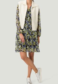 Femme portant une robe à manches longues à motifs, un gilet en fausse fourrure blanc et des baskets argentées, debout avec les jambes croisées devant un fond uni.