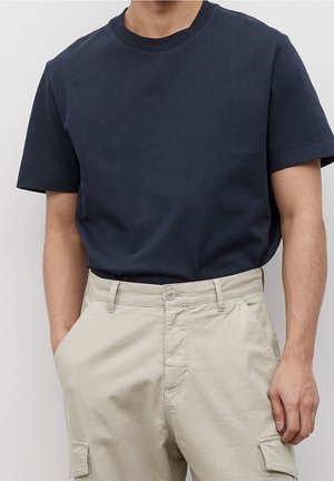 Mann trägt ein schlichtes dunkelblaues Kurzarm-T-Shirt, das in hellbeige Cargo-Hosen gesteckt ist, mit den Händen in den Taschen.