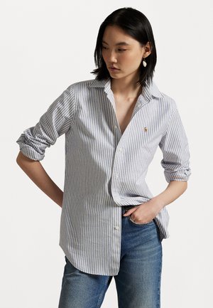 Striped button-up shirt in blue and white cotton; features rolled sleeves, a collar, and a small logo on the chest. Worn with blue jeans.