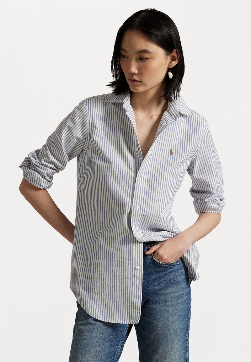 Gestreifte Button-Up-Bluse aus blauem und weißem Baumwollstoff; mit aufgekrempelten Ärmeln, einem Kragen und einem kleinen Logo auf der Brust. Getragen mit blauen Jeans.