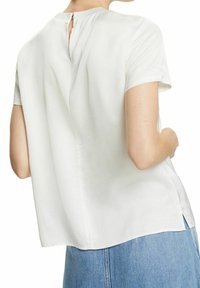 Femme portant un chemisier blanc ample à manches courtes avec une petite fermeture par bouton à l'arrière de l'encolure et un jean en denim bleu clair.