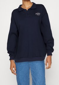 Marinblå tröja med krage, två knappar och långa ärmar. Har "ASPEN"-logotyp i vitt på vänster bröst. Tillverkad av mjukt bomullsmaterial.