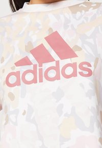 T-shirt bianca a maniche corte con motivo mimetico rosa e un logo "adidas" rosa prominente sul davanti. Realizzata in tessuto leggero.