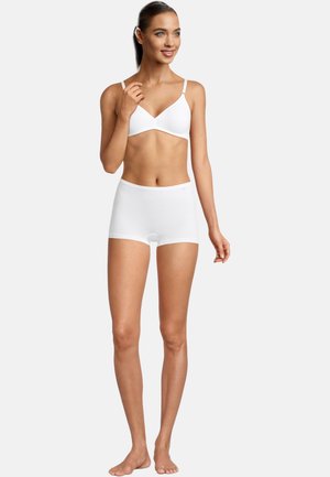 Young woman standing barefoot in white seamless bra and boyshort underwear, smiling and looking to the side on a plain background.