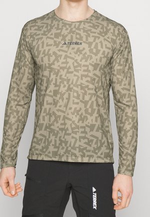 Mann trägt ein langärmeliges, beige-grün gemustertes Hemd mit "TERREX" Logo und schwarze Hose mit weißem "TERREX" Logo am Bein.