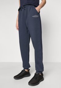 Tamsiai mėlynos laisvalaikio kelnės iš minkštos medvilnės su elastinga juosta, raišteliu, šoninėmis kišenėmis ir balta užrašu "ATHLEISURE".