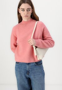 Roza, puhasta pletenina s visokim ovratnikom in rebrastimi manšetami, v kombinaciji z modrimi jeans hlačami in majhno off-white torbico.