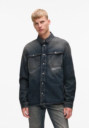 Denim shirt in dark wash, featuring a button-up front, two chest pockets, and a logo detail. Textured fabric with a classic collar design.
