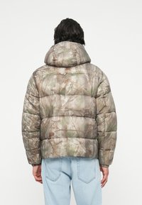 Huzatos kamuflázs pufi dzseki kapucnival, barna és zöld mintákkal, amelyek fák ágaira és lombozatára hasonlítanak.