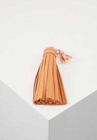 Portachiavi in pelle color cuoio con un piccolo charm a forma di orsacchiotto metallico, adagiato su una superficie bianca inclinata.