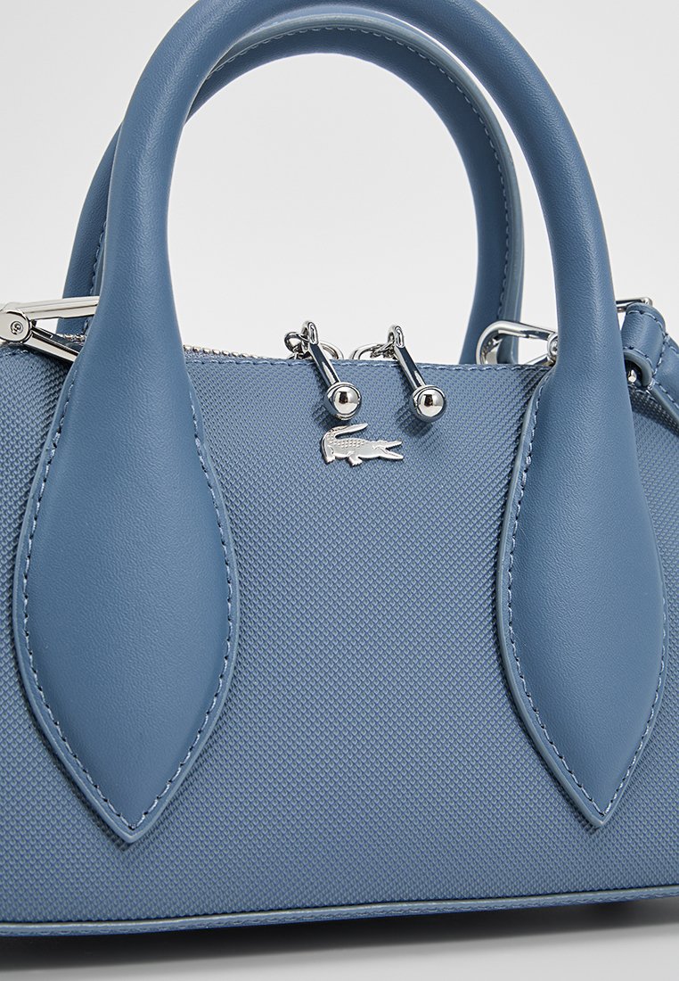 Sac à main en cuir texturé bleu avec des doubles poignées, quincaillerie en argent et accent avec le logo Lacoste. Il présente une forme structurée et des détails lisses.